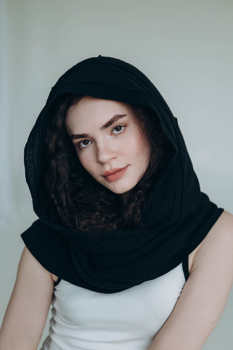May include: A black hooded scarf draped over the shoulders. The scarf is made of a soft, textured fabric. The person is wearing a white tank top. The background is a plain, light color.