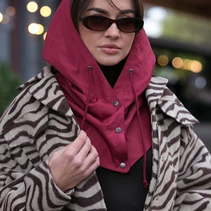 May include: A deep red hooded neck warmer featuring a drawstring closure and three silver-toned snap buttons, worn over a black top and a beige and brown zebra-patterned jacket. The hood is constructed from a smooth, solid-colored fabric.