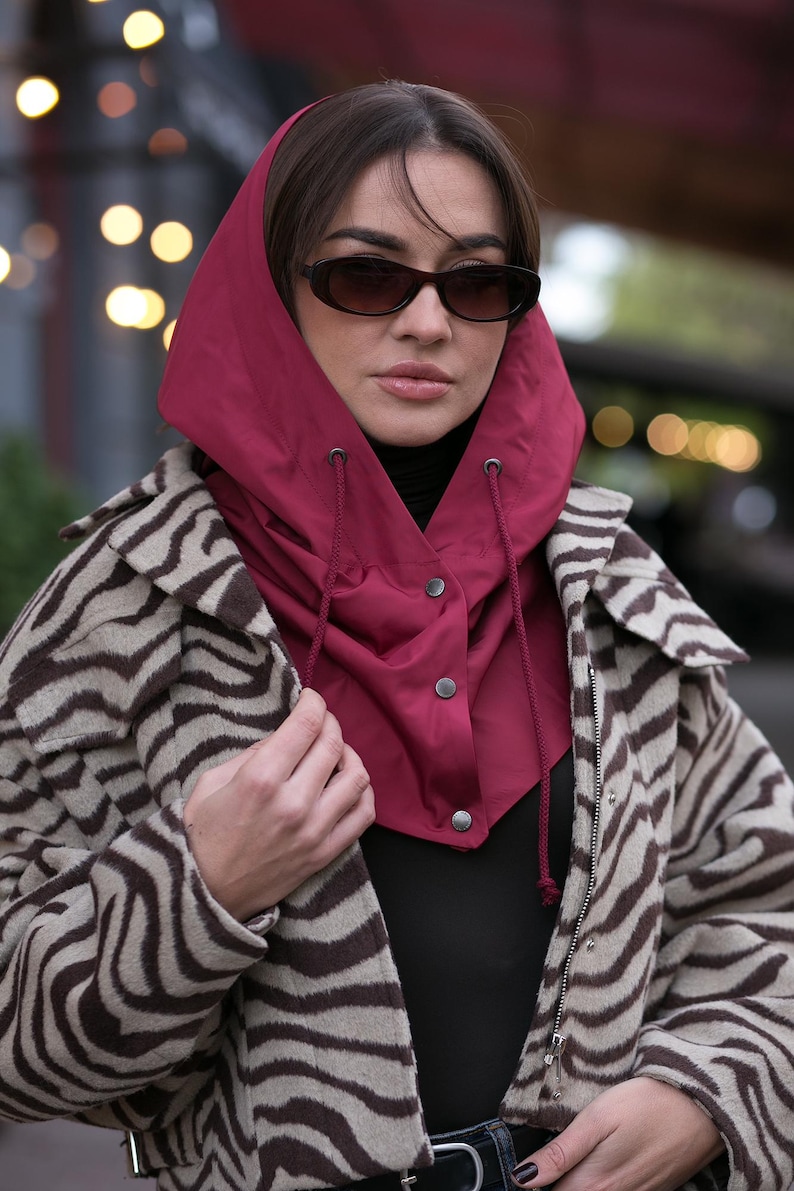 May include: A deep red hooded neck warmer featuring a drawstring closure and three silver-toned snap buttons, worn over a black top and a beige and brown zebra-patterned jacket. The hood is constructed from a smooth, solid-colored fabric.