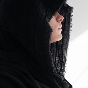 May include: A black hooded scarf made from a textured, crinkled material. The hood frames the face, with a frayed edge adding detail. The scarf is draped around the neck and shoulders, creating a layered effect. The overall aesthetic is dark and mysterious.