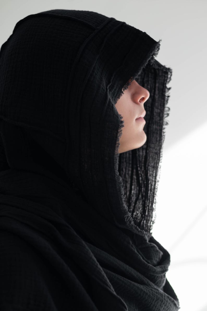May include: A black hooded scarf made from a textured, crinkled material. The hood frames the face, with a frayed edge adding detail. The scarf is draped around the neck and shoulders, creating a layered effect. The overall aesthetic is dark and mysterious.