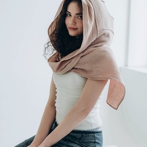 May include: A light pink hooded scarf made of a soft, textured fabric. The scarf is draped over the head and shoulders. The person is wearing a white tank top and gray pants.