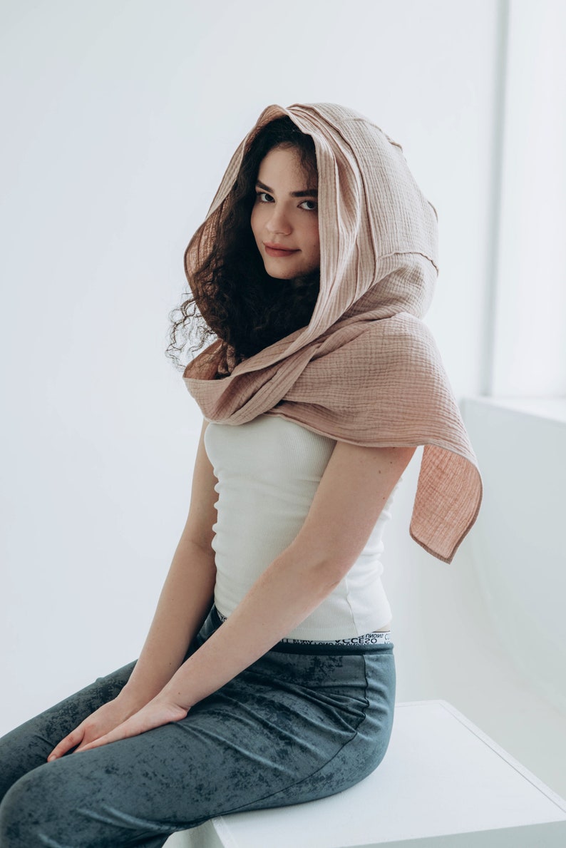 May include: A light pink hooded scarf made of a soft, textured fabric. The scarf is draped over the head and shoulders. The person is wearing a white tank top and gray pants.