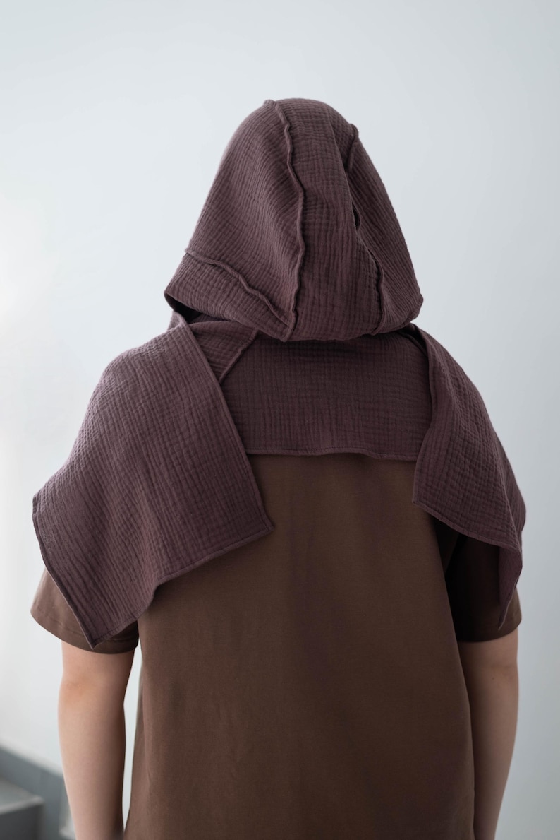 May include: A brown hooded cape and matching short-sleeved shirt. The hood and cape are made of a textured, dark mauve fabric, with visible stitching details. The shirt is a solid, medium brown color, creating a layered effect.
