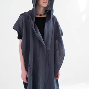May include: A dark grey hooded cape with a draped design. The cape has a long, flowing silhouette and is made of a textured fabric. The model is wearing a black shirt underneath. The hood is also dark grey.