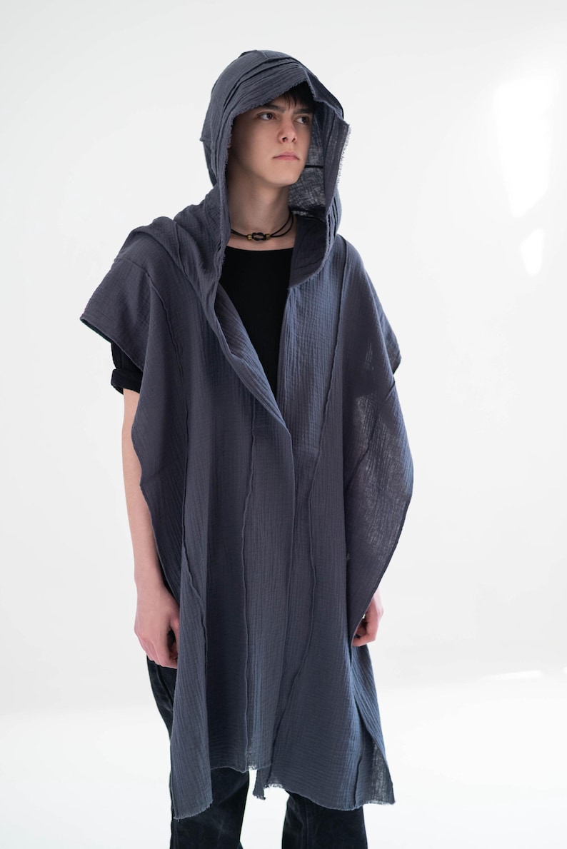 May include: A dark grey hooded cape with a draped design. The cape has a long, flowing silhouette and is made of a textured fabric. The model is wearing a black shirt underneath. The hood is also dark grey.