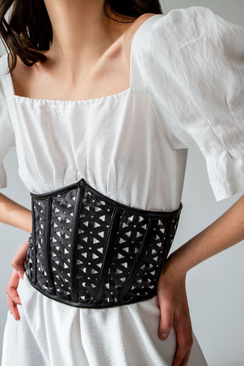 K&ouml;nnte beinhalten: Ein schwarzes Lederkorsett mit geometrischem Cut-Out-Design. Das Korsett wird &uuml;ber einer wei&szlig;en Bluse mit quadratischem Ausschnitt getragen.
