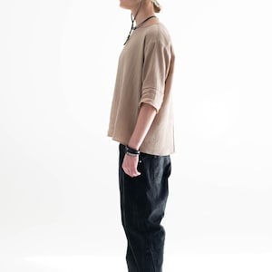May include: A person wearing a tan long-sleeved shirt, black jeans, and a green bucket hat with a frayed brim. The outfit includes a black bracelet and necklace. The person is wearing grey trainers.