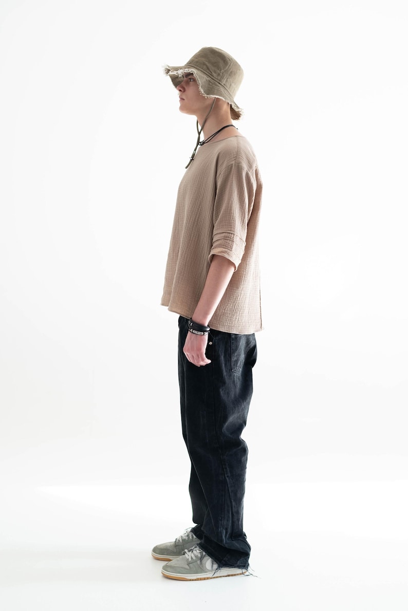 May include: A person wearing a tan long-sleeved shirt, black jeans, and a green bucket hat with a frayed brim. The outfit includes a black bracelet and necklace. The person is wearing grey trainers.