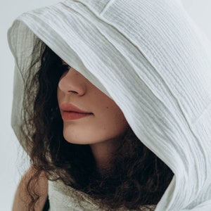 May include: A white hooded scarf made of a textured fabric. The hood is draped over the head and shoulders, partially obscuring the face. The fabric has a subtle, ribbed pattern. The overall aesthetic is soft and minimalist.