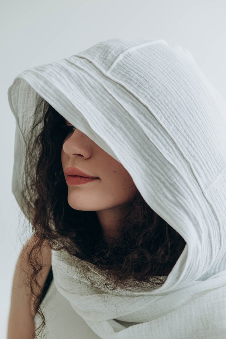 May include: A white hooded scarf made of a textured fabric. The hood is draped over the head and shoulders, partially obscuring the face. The fabric has a subtle, ribbed pattern. The overall aesthetic is soft and minimalist.