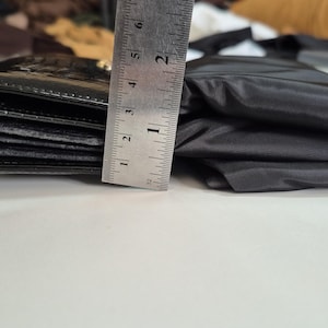 May include: A black wallet with a textured surface next to a folded black fabric item. A metal ruler is positioned vertically against the side of the wallet, showing a height measurement of approximately 2.25 inches for the wallet's thickness.