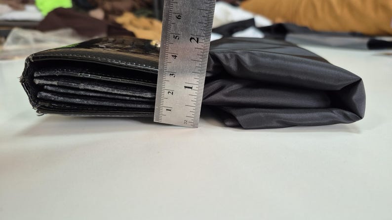 May include: A black wallet with a textured surface next to a folded black fabric item. A metal ruler is positioned vertically against the side of the wallet, showing a height measurement of approximately 2.25 inches for the wallet's thickness.