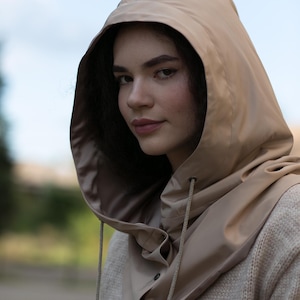 May include: A beige hooded cowl made of smooth, water-resistant material featuring a drawstring closure with matching cords and a buttoned front placket, worn over a textured, light-colored knit sweater with visible buttons.