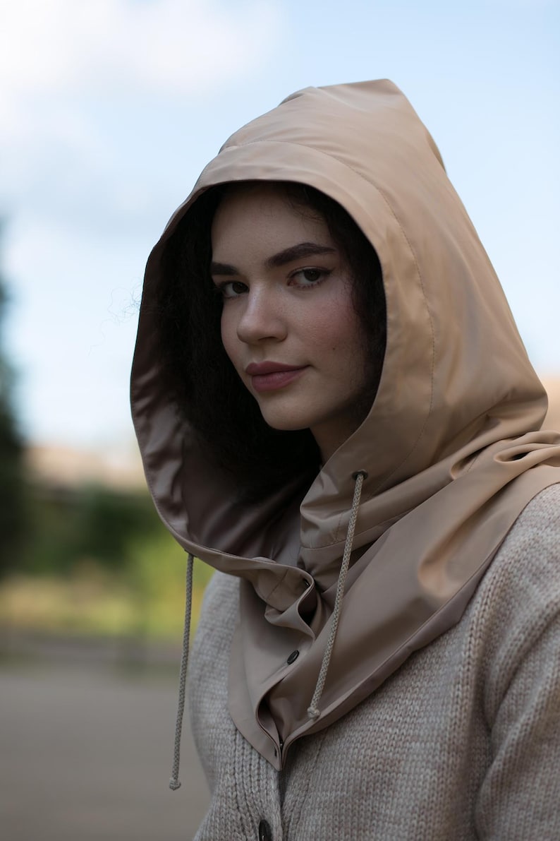 May include: A beige hooded cowl made of smooth, water-resistant material featuring a drawstring closure with matching cords and a buttoned front placket, worn over a textured, light-colored knit sweater with visible buttons.