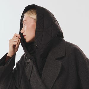 May include: A dark charcoal gray wool-blend coat featuring a black quilted hood with a geometric stitched pattern and black drawstring cords. The coat has wide lapels and a structured silhouette, providing a layered look with the attached hood.