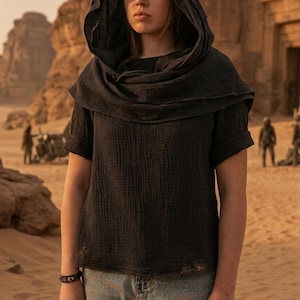 May include: A dark gray hooded top with short sleeves and a textured appearance. The top is paired with light blue denim jeans. The background features a desert landscape with rock formations and figures in the distance.