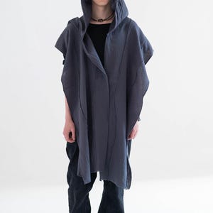 May include: A dark grey hooded cape with vertical seam detailing. The cape is worn over a black top and dark blue jeans. The person is also wearing a black necklace and grey trainers.