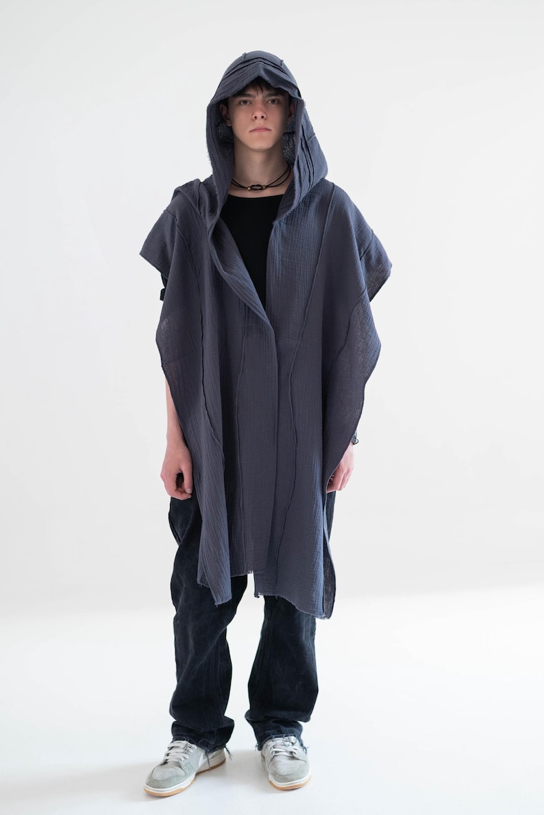 May include: A dark grey hooded cape with vertical seam detailing. The cape is worn over a black top and dark blue jeans. The person is also wearing a black necklace and grey trainers.