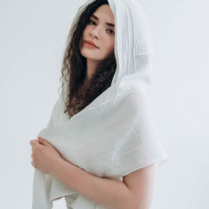 May include: A white hooded scarf made of a soft, textured fabric. The scarf is draped over the shoulders and head, creating a flowing, elegant look. The fabric appears to be lightweight and breathable, perfect for layering.