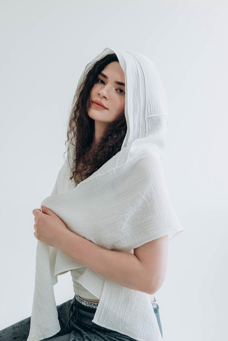 May include: A white hooded scarf made of a soft, textured fabric. The scarf is draped over the shoulders and head, creating a flowing, elegant look. The fabric appears to be lightweight and breathable, perfect for layering.