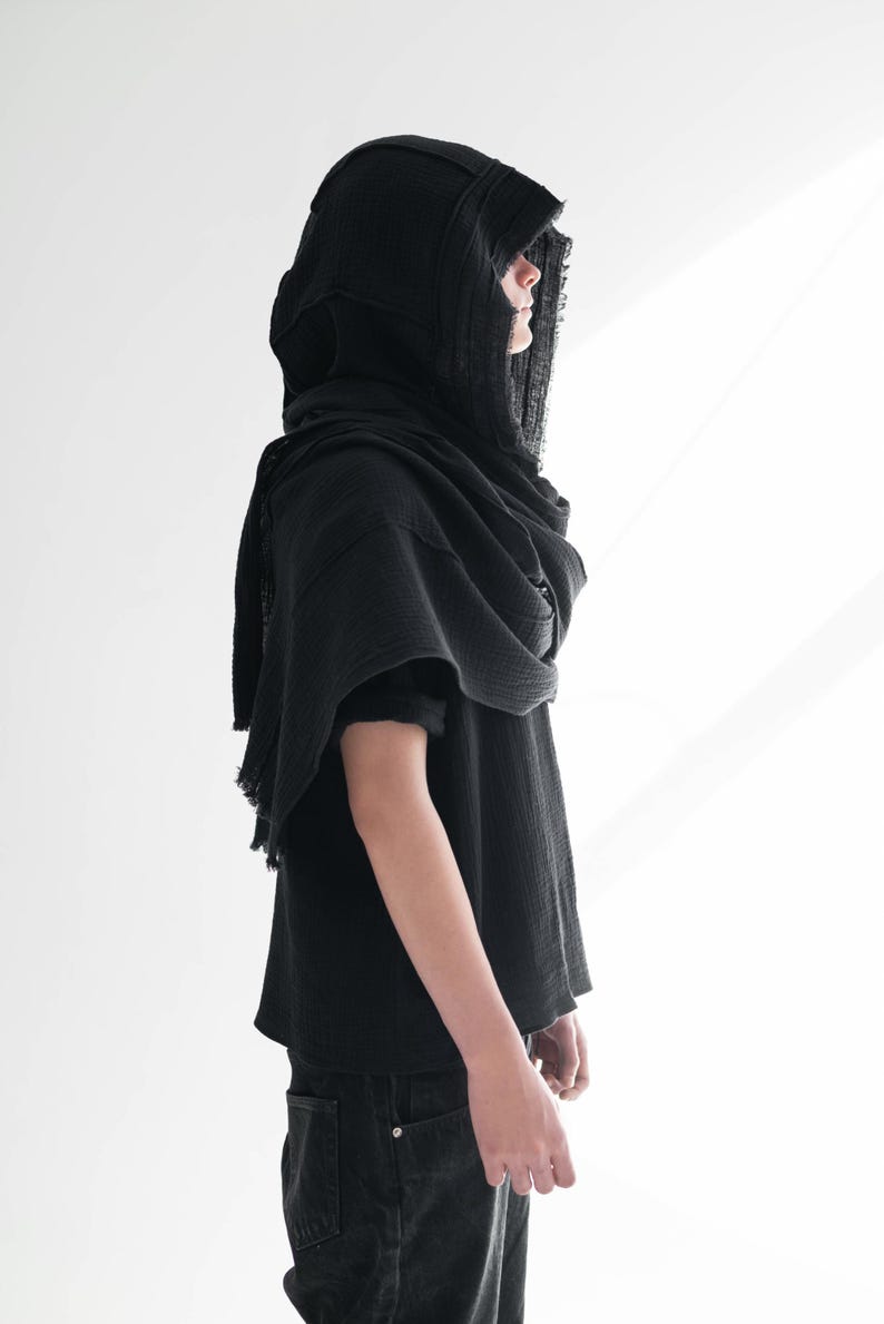 May include: A black hooded scarf and top. The hood is draped over the shoulders, and the top has short sleeves. The fabric appears to be a lightweight, textured material. The person is wearing black jeans.
