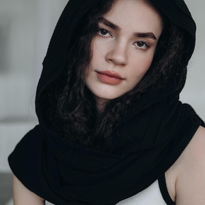 May include: A person wearing a black hood and a white tank top. The hood is draped over the head and shoulders, creating a dark frame around the face. The person has dark, curly hair and is looking at the camera.