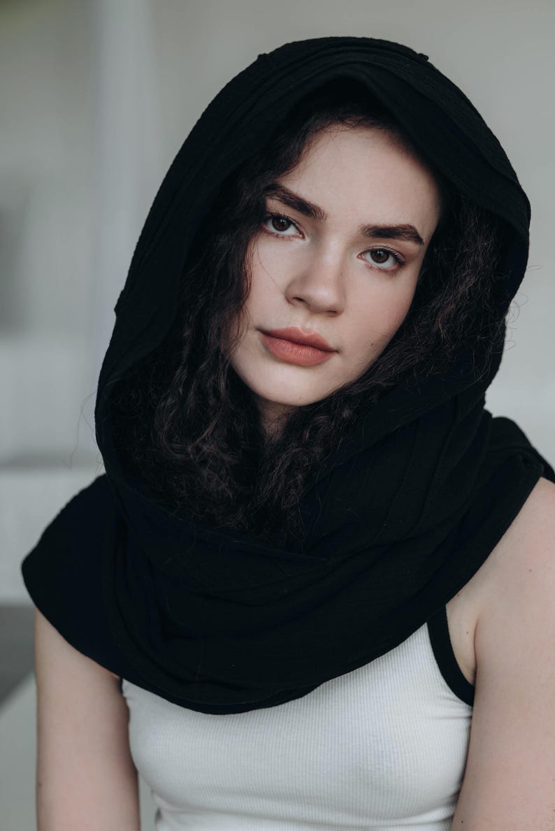 May include: A person wearing a black hood and a white tank top. The hood is draped over the head and shoulders, creating a dark frame around the face. The person has dark, curly hair and is looking at the camera.