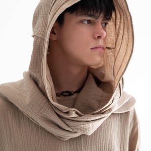 May include: A beige hooded shirt with a textured, ribbed fabric. The hood is draped around the neck and shoulders. A black necklace with a small pendant is visible. The shirt is a neutral color, suitable for various styles.