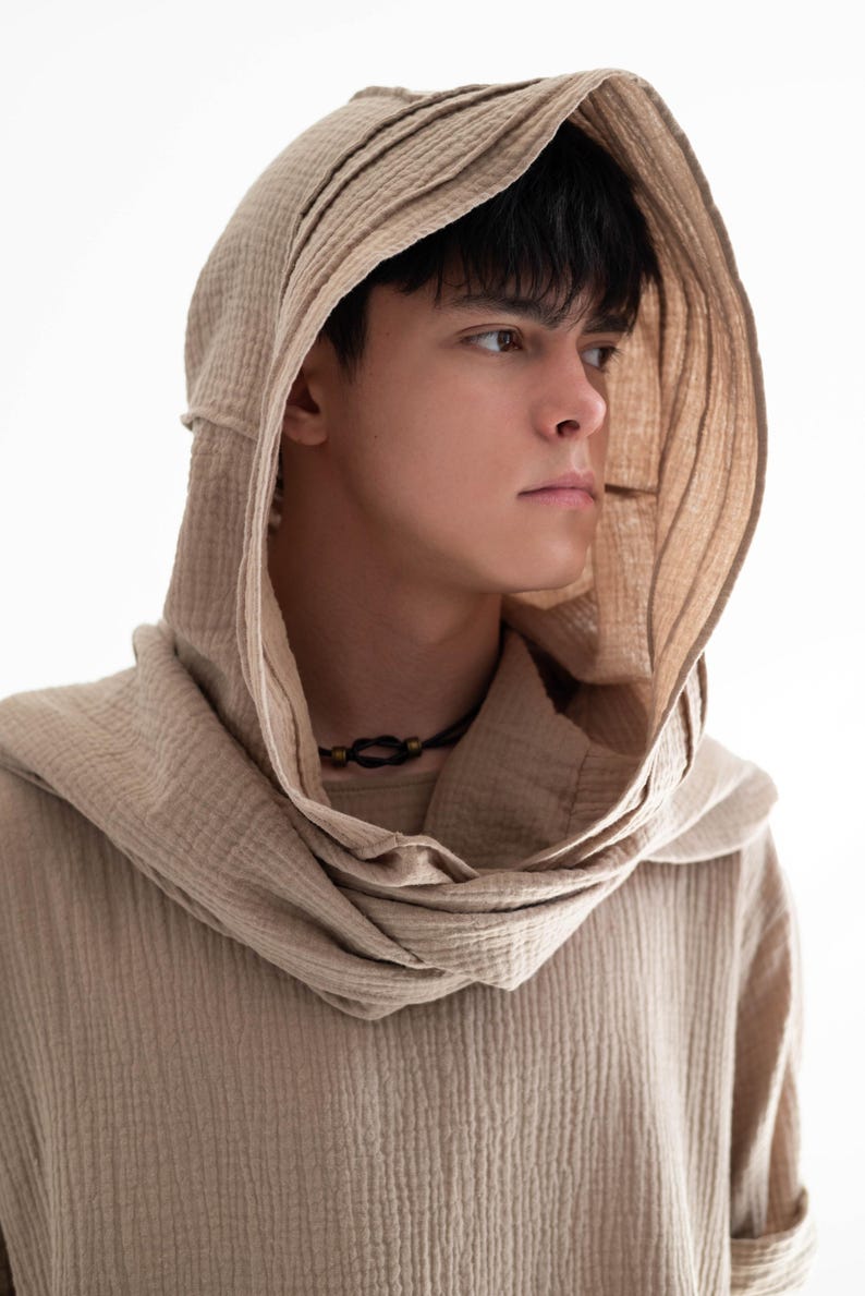 May include: A beige hooded shirt with a textured, ribbed fabric. The hood is draped around the neck and shoulders. A black necklace with a small pendant is visible. The shirt is a neutral color, suitable for various styles.