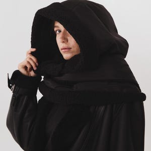 May include: A black hooded jacket featuring a thick, textured black faux shearling lining along the hood edge and front opening. The jacket is constructed from a smooth, matte black fabric and includes a wide collar with matching black trim.