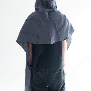 May include: A dark grey hooded cape with a layered design, worn over a black top and dark grey jeans. The cape features visible stitching and a draped silhouette, creating a unique, textured look. The person is facing away from the camera.