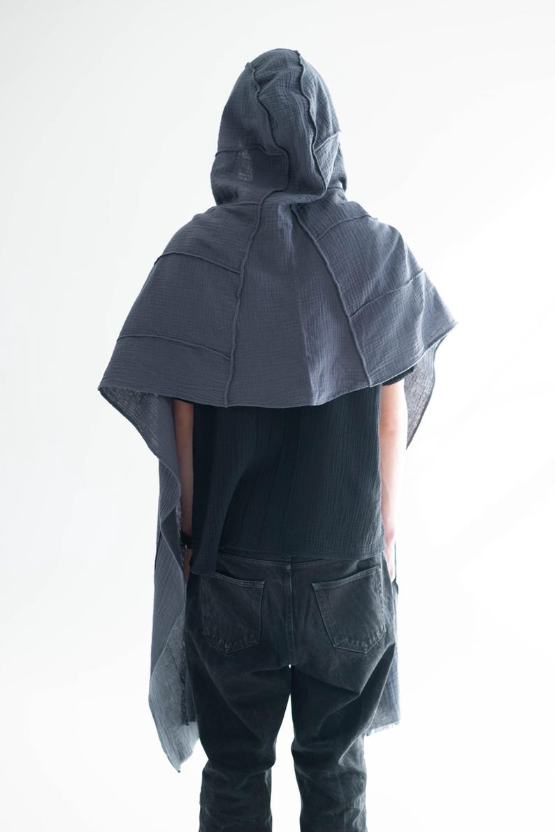 May include: A dark grey hooded cape with a layered design, worn over a black top and dark grey jeans. The cape features visible stitching and a draped silhouette, creating a unique, textured look. The person is facing away from the camera.