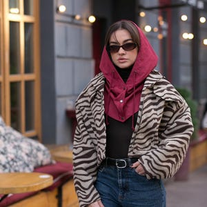 May include: A cropped jacket with a brown and beige zebra print pattern, worn over a black top and blue jeans. A deep red hooded balaclava with a front button closure and drawstring ties is layered underneath the jacket. Dark oval sunglasses.