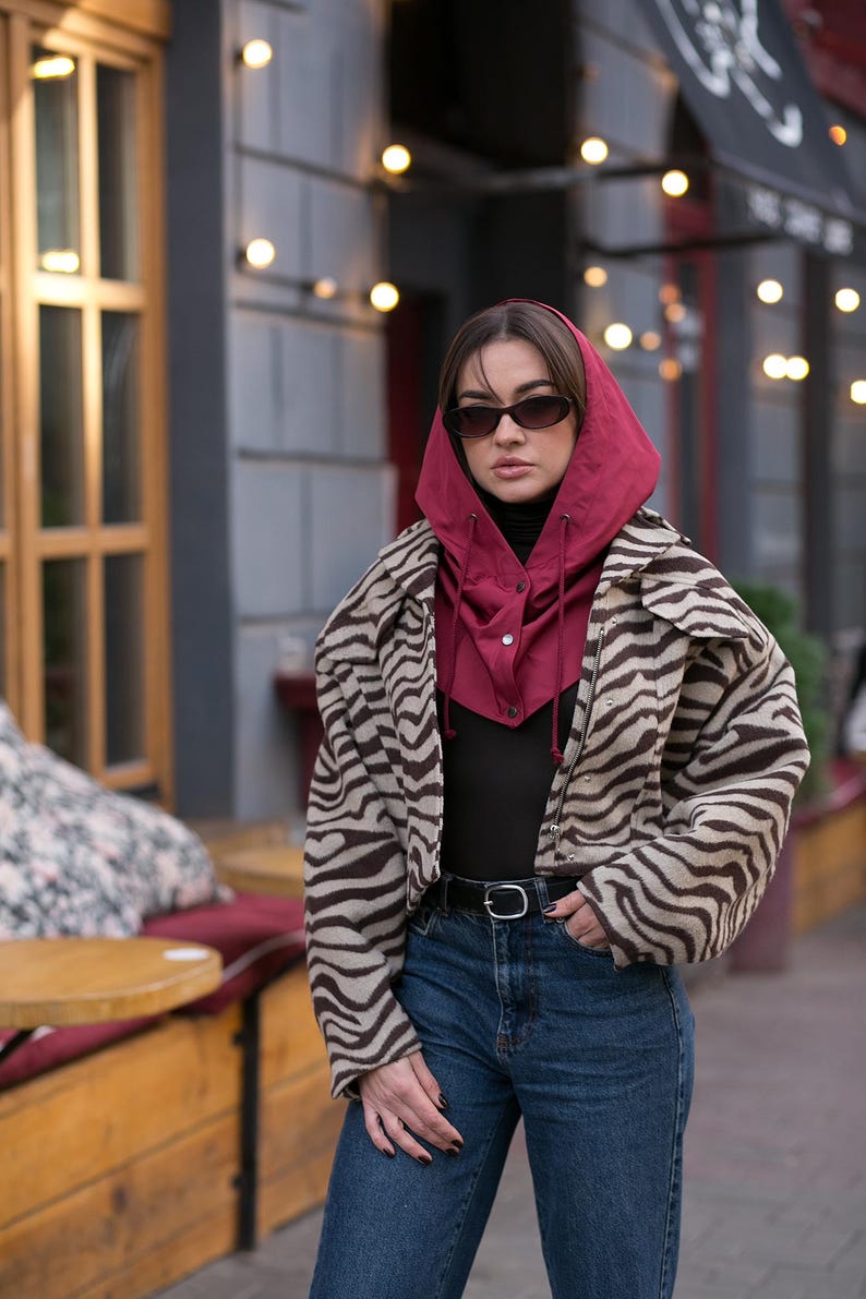 May include: A cropped jacket with a brown and beige zebra print pattern, worn over a black top and blue jeans. A deep red hooded balaclava with a front button closure and drawstring ties is layered underneath the jacket. Dark oval sunglasses.