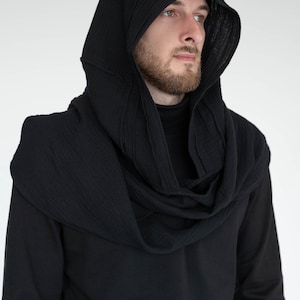 May include: Black hooded scarf and cowl combination. The hood has a structured design with vertical stitching, and the cowl wraps around the neck. The garment is made of a textured fabric and is worn over a black long-sleeve shirt. A modern, stylish accessory.