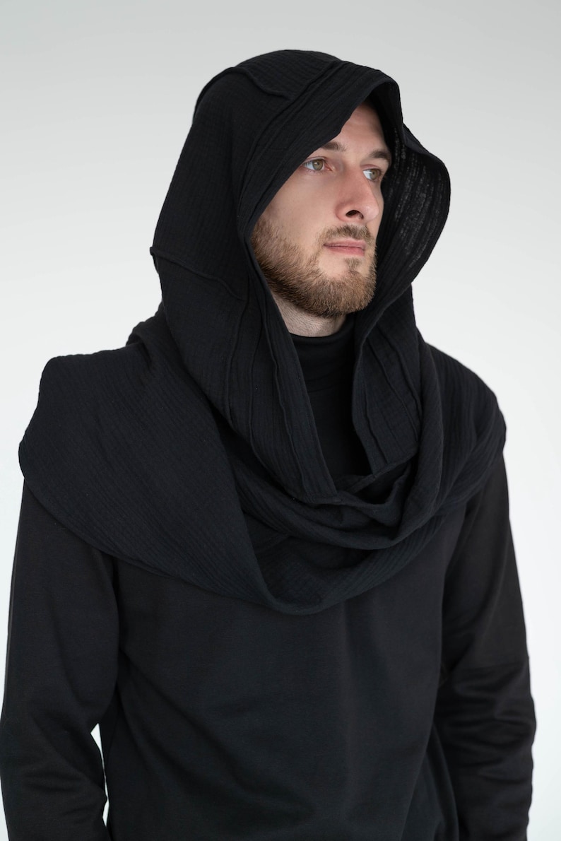May include: Black hooded scarf and cowl combination. The hood has a structured design with vertical stitching, and the cowl wraps around the neck. The garment is made of a textured fabric and is worn over a black long-sleeve shirt. A modern, stylish accessory.