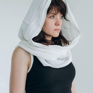 May include: A white hooded scarf draped over a black tank top. The scarf is made of a soft, textured fabric and frames the face. The overall look is simple and elegant, suitable for various occasions.