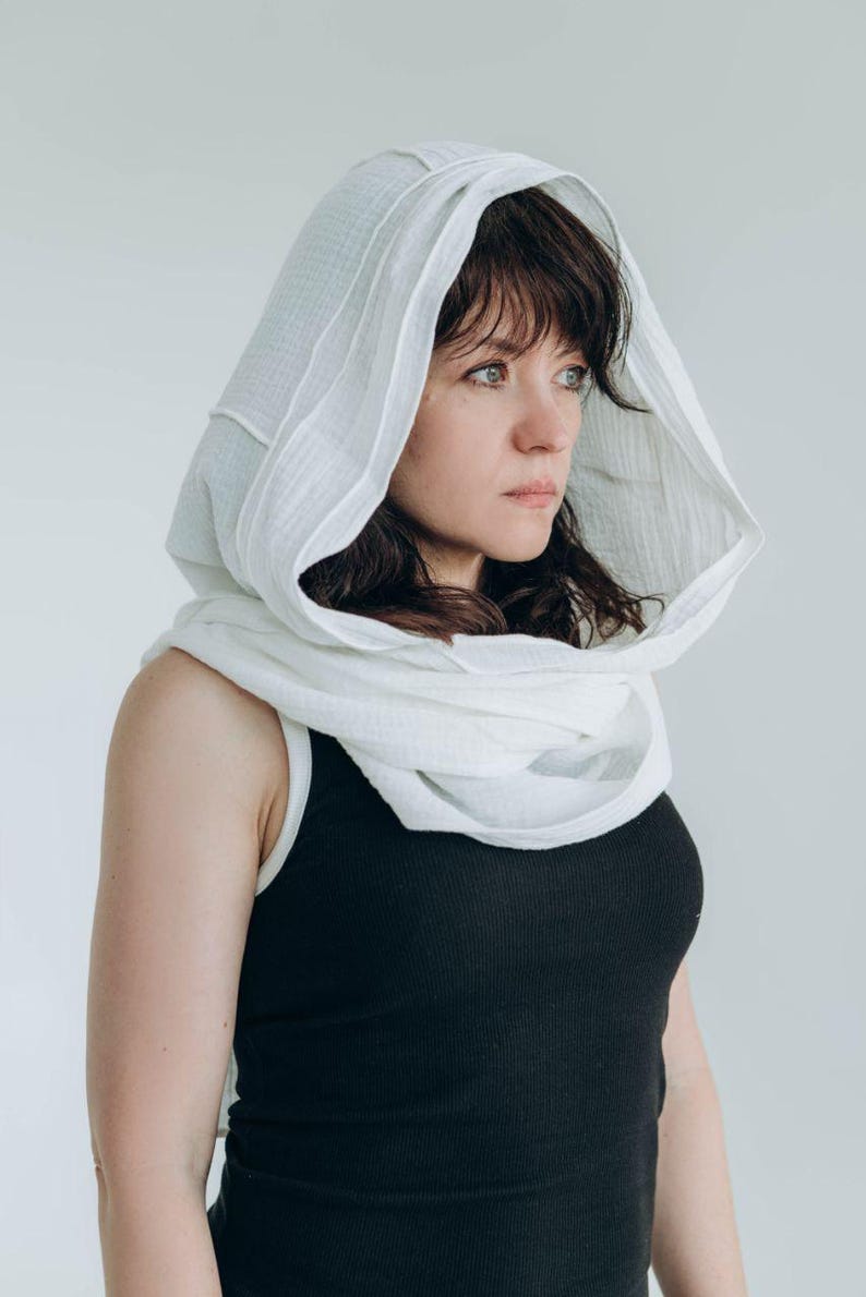 May include: A white hooded scarf draped over a black tank top. The scarf is made of a soft, textured fabric and frames the face. The overall look is simple and elegant, suitable for various occasions.