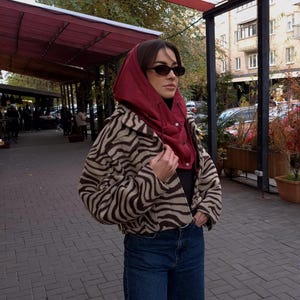 May include: A cropped jacket with a brown and beige zebra print pattern, worn over a dark top and a deep red hooded balaclava. The outfit is completed with dark blue denim jeans and small, dark-tinted oval sunglasses, creating a layered look.