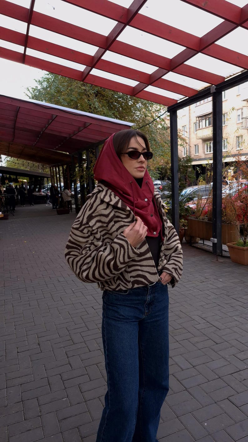 May include: A cropped jacket with a brown and beige zebra print pattern, worn over a dark top and a deep red hooded balaclava. The outfit is completed with dark blue denim jeans and small, dark-tinted oval sunglasses, creating a layered look.