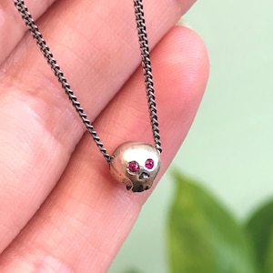 Könnte beinhalten: Eine silberne Totenkopf-Anhängerkette mit roten Edelsteinaugen an einer schwarzen Kette.