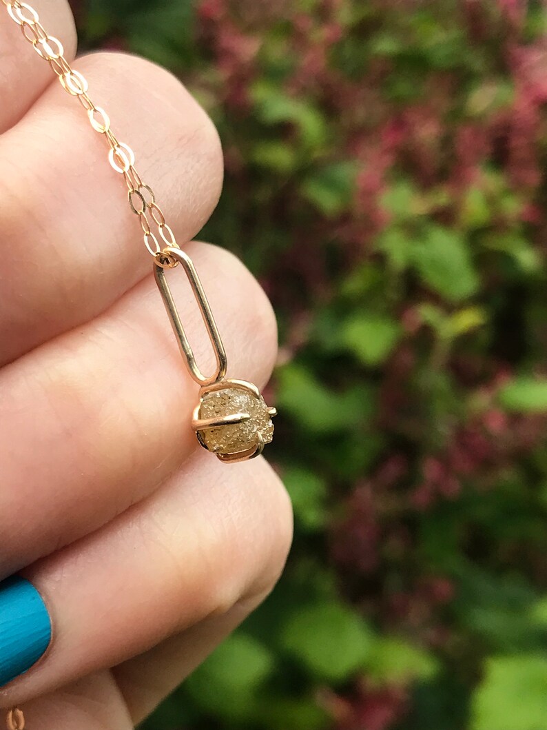 May include: A gold chain necklace with a small, round, raw diamond pendant. The diamond is set in a gold cage.