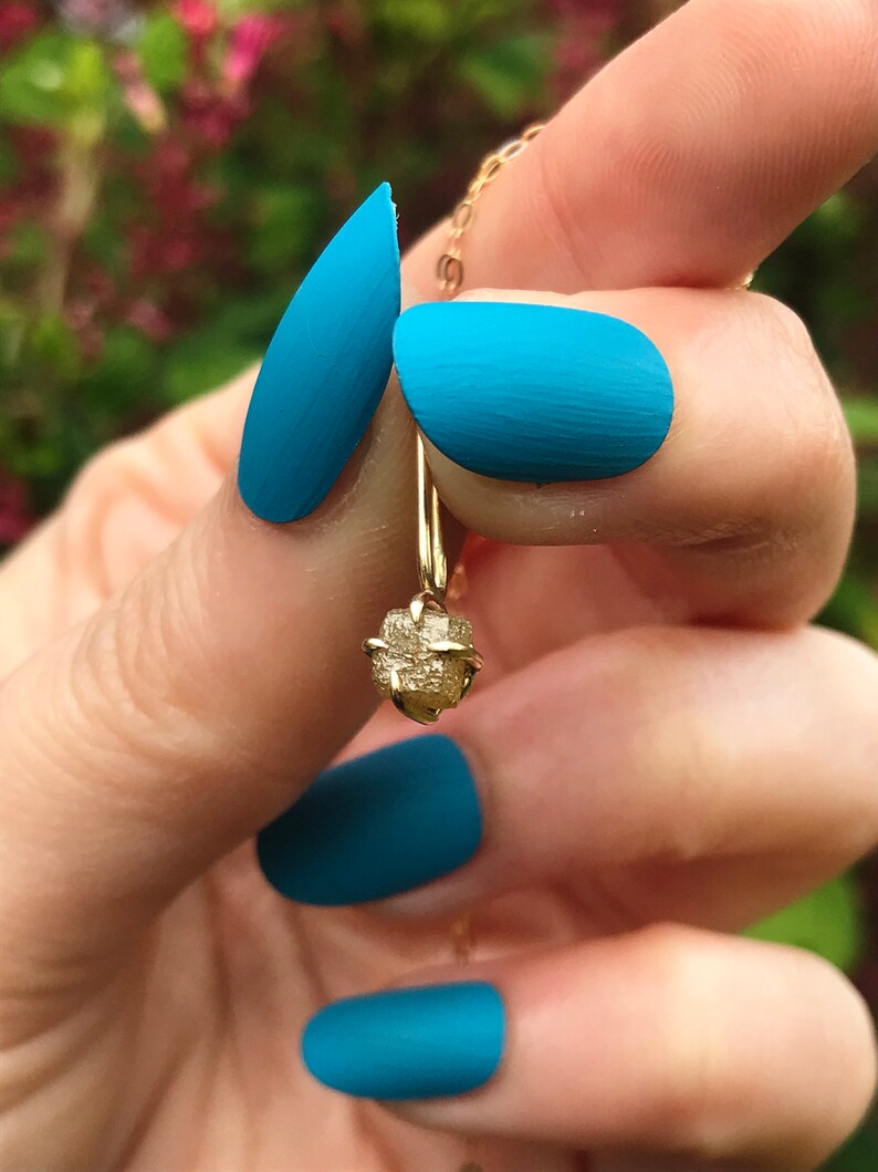 May include: A gold pendant necklace with a rough, raw diamond-shaped stone. The necklace is hanging from a person's finger with blue nail polish.