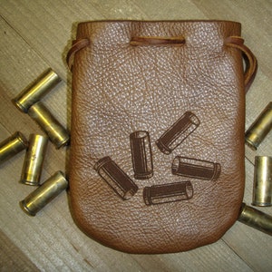 May include: A brown leather pouch with a drawstring closure, surrounded by several brass bullet casings. The pouch has a textured surface and a decorative design of bullet casings. The items are arranged on a wooden surface.