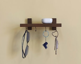 May include: A dark brown wooden wall mounted key rack with four hooks. A pair of glasses hangs on one hook, a small white rectangular box sits on the shelf, and three keychains hang from the other hooks.