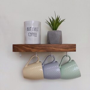May include: A wooden shelf with three hooks holding three ceramic mugs. The mugs are beige, gray, and green. A white ceramic mug with the text "But First Coffee" is sitting on the shelf. A small potted plant is also on the shelf.