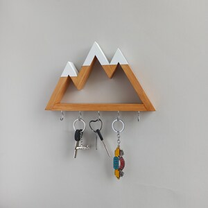 May include: Wooden key holder shaped like a mountain range with white peaks. The holder has five hooks for hanging keys. There are three keys hanging on the hooks.
