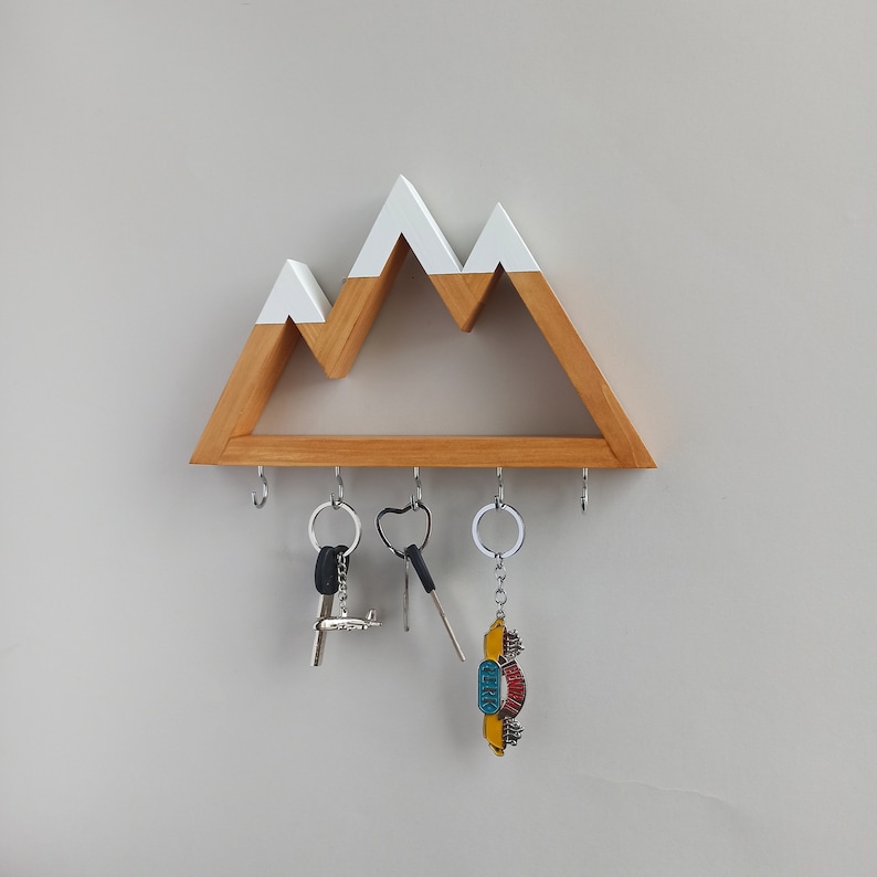 May include: Wooden key holder shaped like a mountain range with white peaks. The holder has five hooks for hanging keys. There are three keys hanging on the hooks.