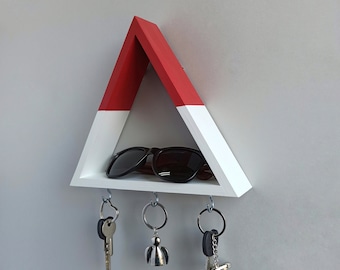 May include: A white and red triangular key holder with three hooks and a shelf for sunglasses. The key holder is mounted on a white wall.